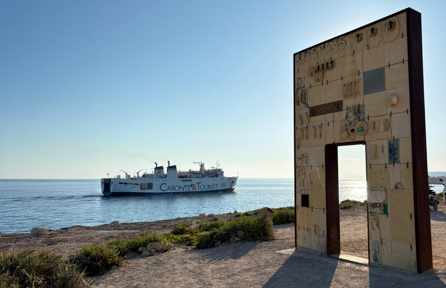A Lampedusa la Porta d’Europa, il monumento dedicato alla memoria dei migranti che hanno perso la vita in mare, e sullo sfondo uno dei traghetti che trasferiscono i migranti in Sicilia, a Porto Empedocle